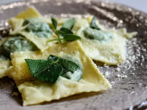 Ravioli ricotta e spinaci, conditi con burro fuso e foglie di salvia fresca, spolverati di parmigiano grattugiato su un piatto elegante.