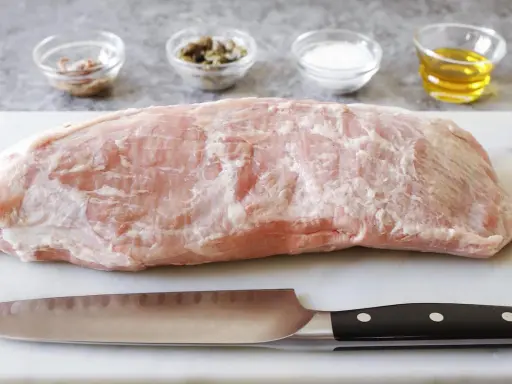 Un pezzo di girello di vitello crudo, pronto per essere cucinato, con coltello e ciotoline di condimenti sullo sfondo.