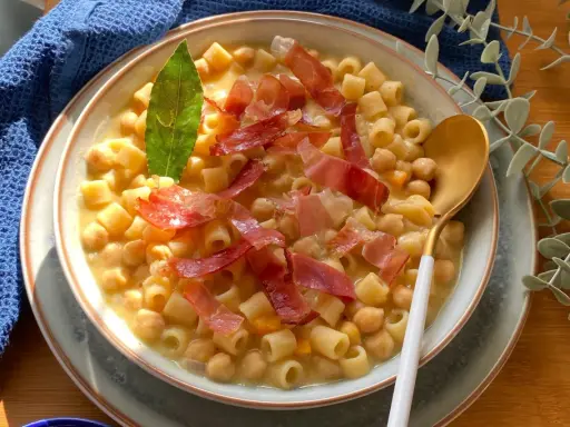Un piatto fumante di pasta e ceci, arricchito da fette di prosciutto crudo e una foglia di alloro.