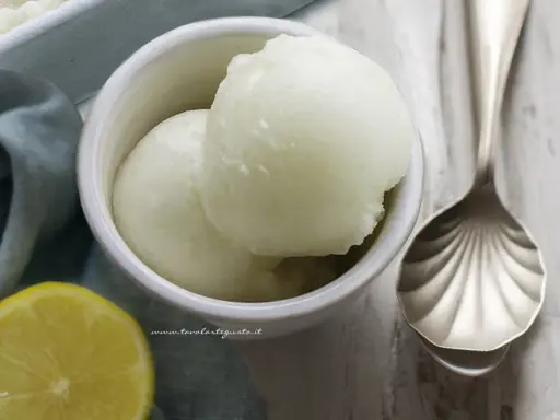 Due palline di sorbetto al limone senza albume in una ciotola bianca, con una fetta di limone e un cucchiaio decorato.
