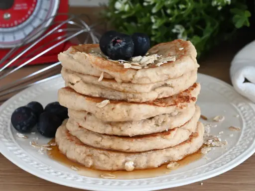 Pila di pancake con farina di avena, guarniti con mirtilli e fiocchi d'avena, su un piatto bianco con sciroppo d'acero.