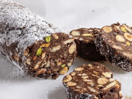 Salame di cioccolato senza uova, ricco di nocciole, mandorle e pistacchi, spolverato di zucchero a velo.