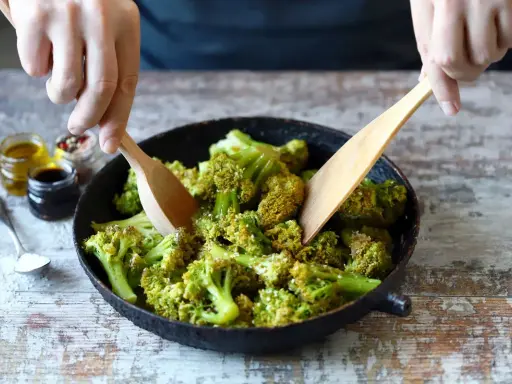 Mani che mescolano broccoli in padella con due palette di legno. Sullo sfondo, piccoli barattoli con olio, aceto balsamico e spezie.