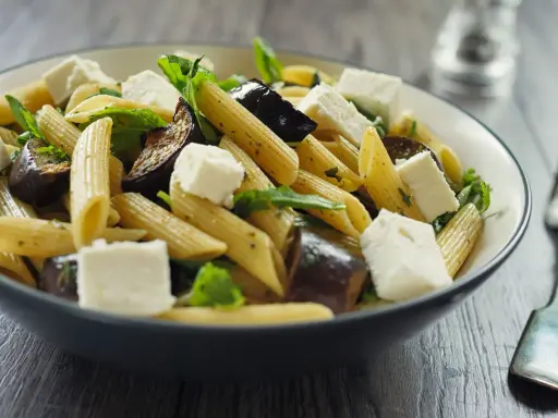 Piatto di pasta con melanzane in bianco, cubetti di formaggio fresco e foglie verdi, servito in una ciotola scura.
