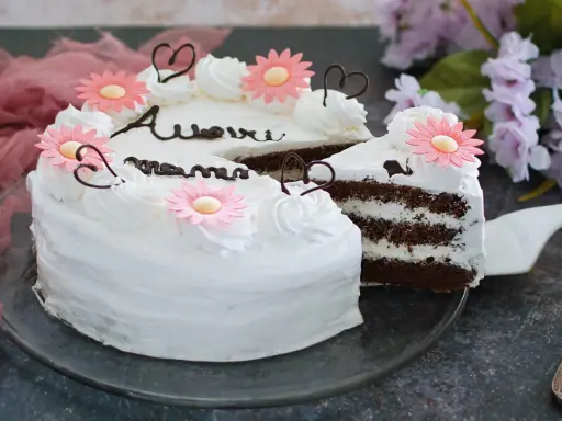 Una torta festa della mamma con glassa bianca, fiori rosa e cuori di cioccolato. La fetta tagliata rivela strati di torta al cioccolato e crema.