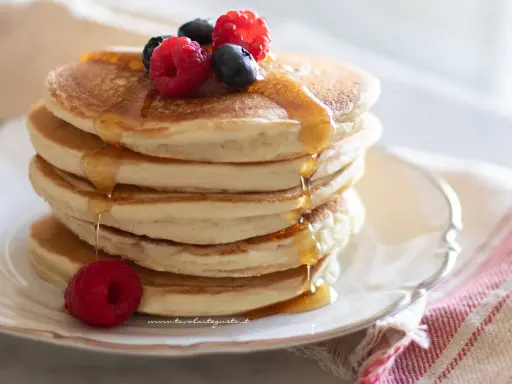 Pancakes senza glutine dorati, impilati e inzuppati di sciroppo, guarniti con lamponi e mirtilli freschi.