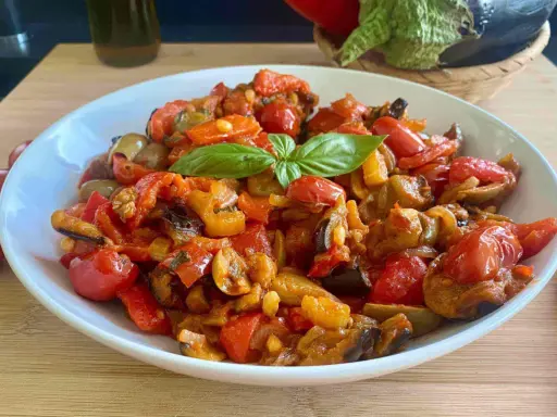 Caponata siciliana ricetta della nonna: melanzane, pomodori, peperoni e basilico in un piatto bianco.