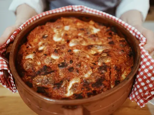 Un invitante pasta al forno appena sfornata, con crosta dorata e ripieno filante, tenuta con un canovaccio a quadretti rossi.