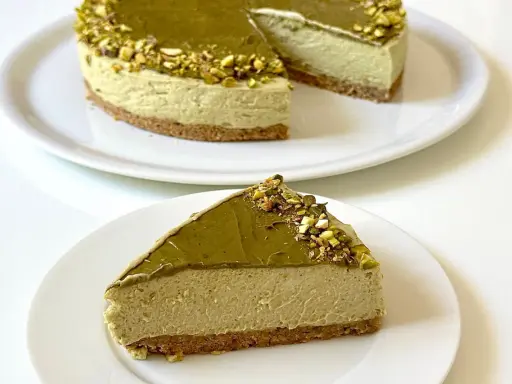 Una fetta di goloso cheesecake al pistacchio, con topping e granella di pistacchi, su un piatto bianco.
