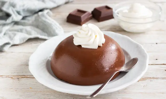 Budino al cioccolato liscio e lucido, guarnito con panna montata, su un piatto bianco. Accanto, due quadratini di cioccolato e una ciotolina di panna.