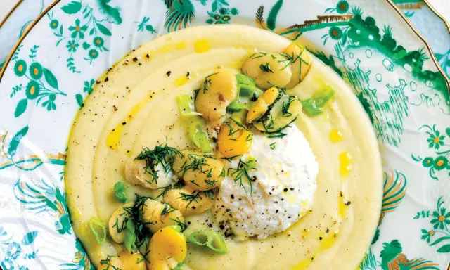 Un cremoso macco di fave fresche, guarnito con ricotta, fave intere, cipollotto e aneto, servito su un piatto decorato.