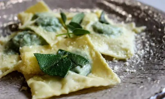 Ravioli ricotta e spinaci, conditi con burro fuso e foglie di salvia fresca, spolverati di parmigiano grattugiato su un piatto elegante.