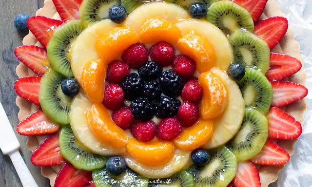 Torta alla frutta coloratissima, con fragole, kiwi, mandarini, lamponi e mirtilli disposti a raggiera su una base croccante.