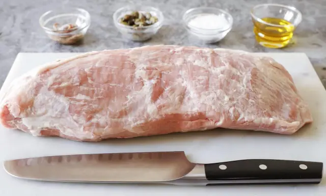 Un pezzo di girello di vitello crudo, pronto per essere cucinato, con coltello e ciotoline di condimenti sullo sfondo.