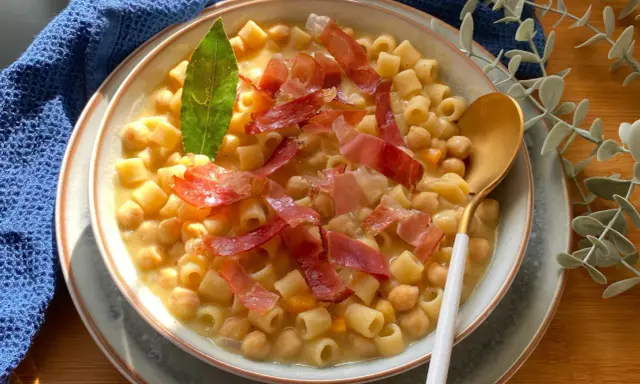 Un piatto fumante di pasta e ceci, arricchito da fette di prosciutto crudo e una foglia di alloro.