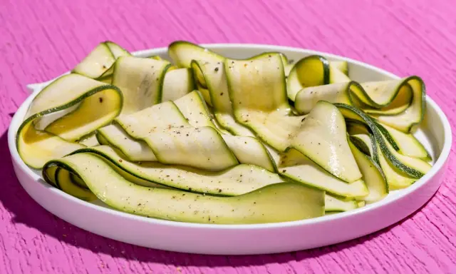 Fettine sottili di zucchine marinate crude, condite con pepe nero, disposte a spirale in un piatto bianco su un tavolo rosa acceso.