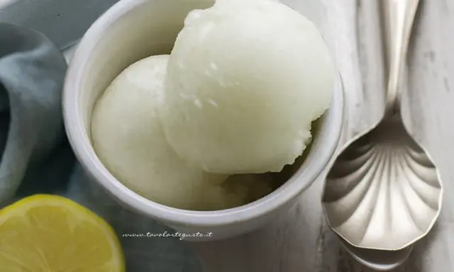 Due palline di sorbetto al limone senza albume in una ciotola bianca, con una fetta di limone e un cucchiaio decorato.
