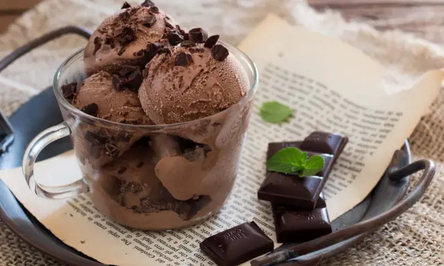 Coppa di gelato al cioccolato con scaglie di cioccolato fondente e foglioline di menta, su un vassoio arrugginito con un libro aperto.