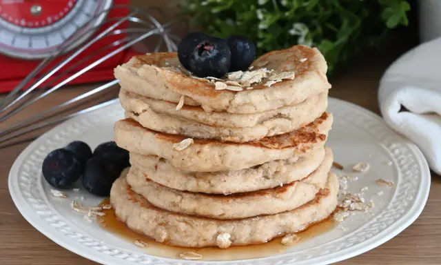 Pila di pancake con farina di avena, guarniti con mirtilli e fiocchi d'avena, su un piatto bianco con sciroppo d'acero.