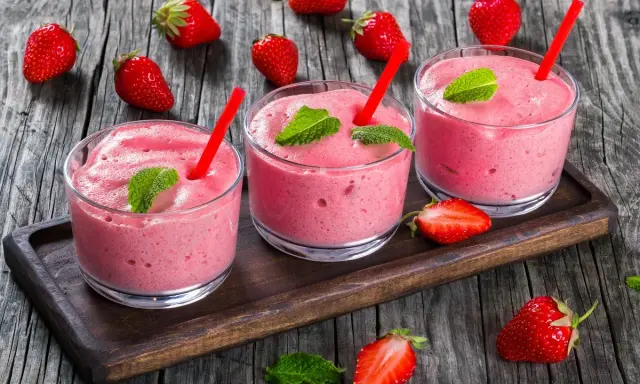 Tre bicchieri di spumone alle fragole bimby, guarniti con menta e fragole fresche, su un vassoio di legno.