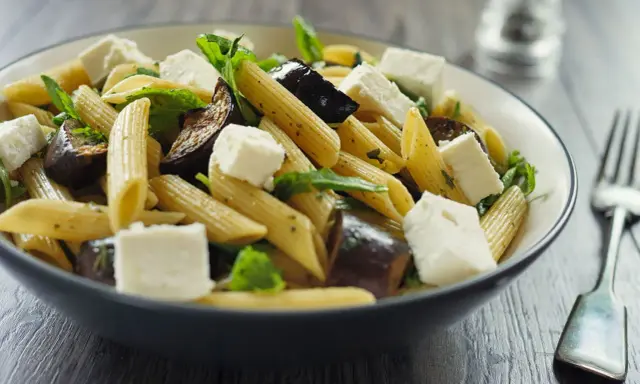 Piatto di pasta con melanzane in bianco, cubetti di formaggio fresco e foglie verdi, servito in una ciotola scura.
