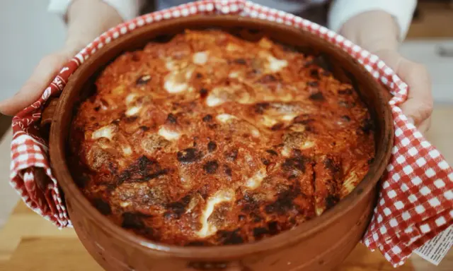 Un invitante pasta al forno appena sfornata, con crosta dorata e ripieno filante, tenuta con un canovaccio a quadretti rossi.