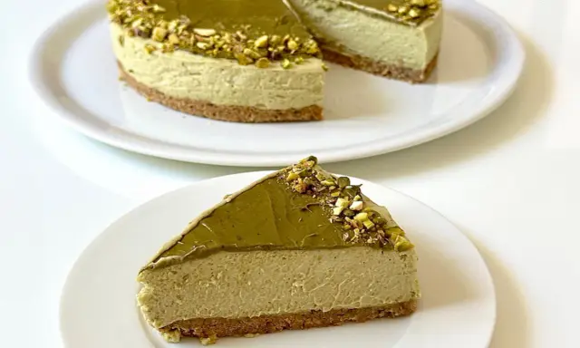 Una fetta di goloso cheesecake al pistacchio, con topping e granella di pistacchi, su un piatto bianco.