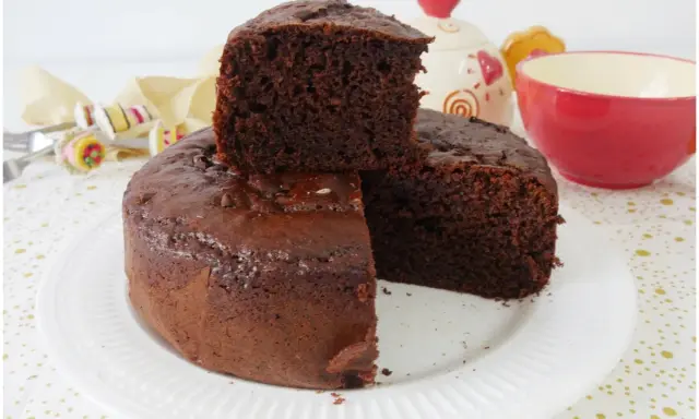 Una fetta di torta yogurt e cioccolato, soffice e golosa, è appoggiata su una torta intera. Sullo sfondo, una tazza rossa e un barattolo decorato.
