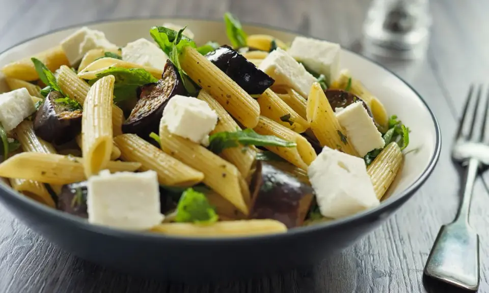 Piatto di pasta con melanzane in bianco, cubetti di formaggio fresco e foglie verdi, servito in una ciotola scura.