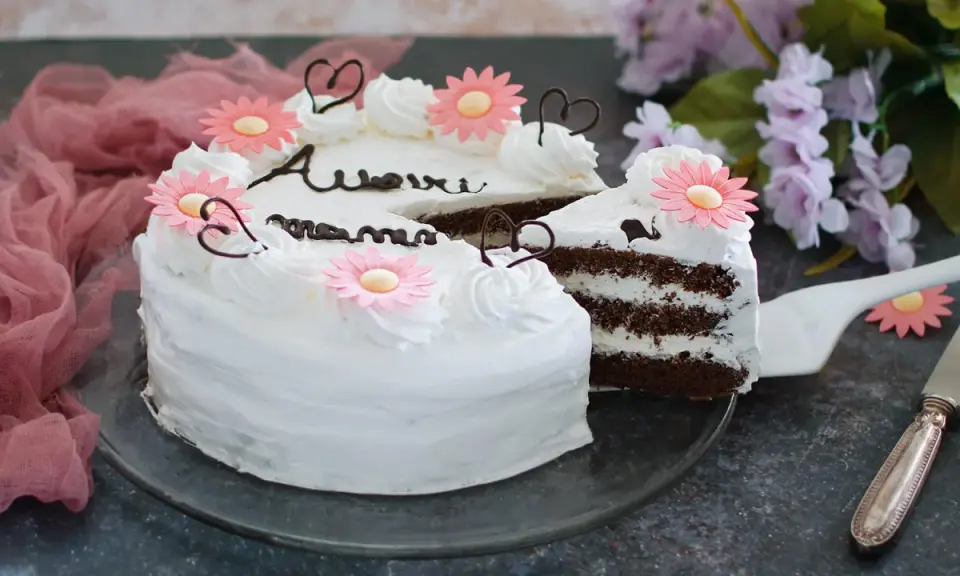 Una torta festa della mamma con glassa bianca, fiori rosa e cuori di cioccolato. La fetta tagliata rivela strati di torta al cioccolato e crema.