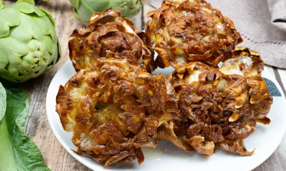 Carciofi alla giudia dorati e croccanti su un piatto bianco, con carciofi freschi sullo sfondo.