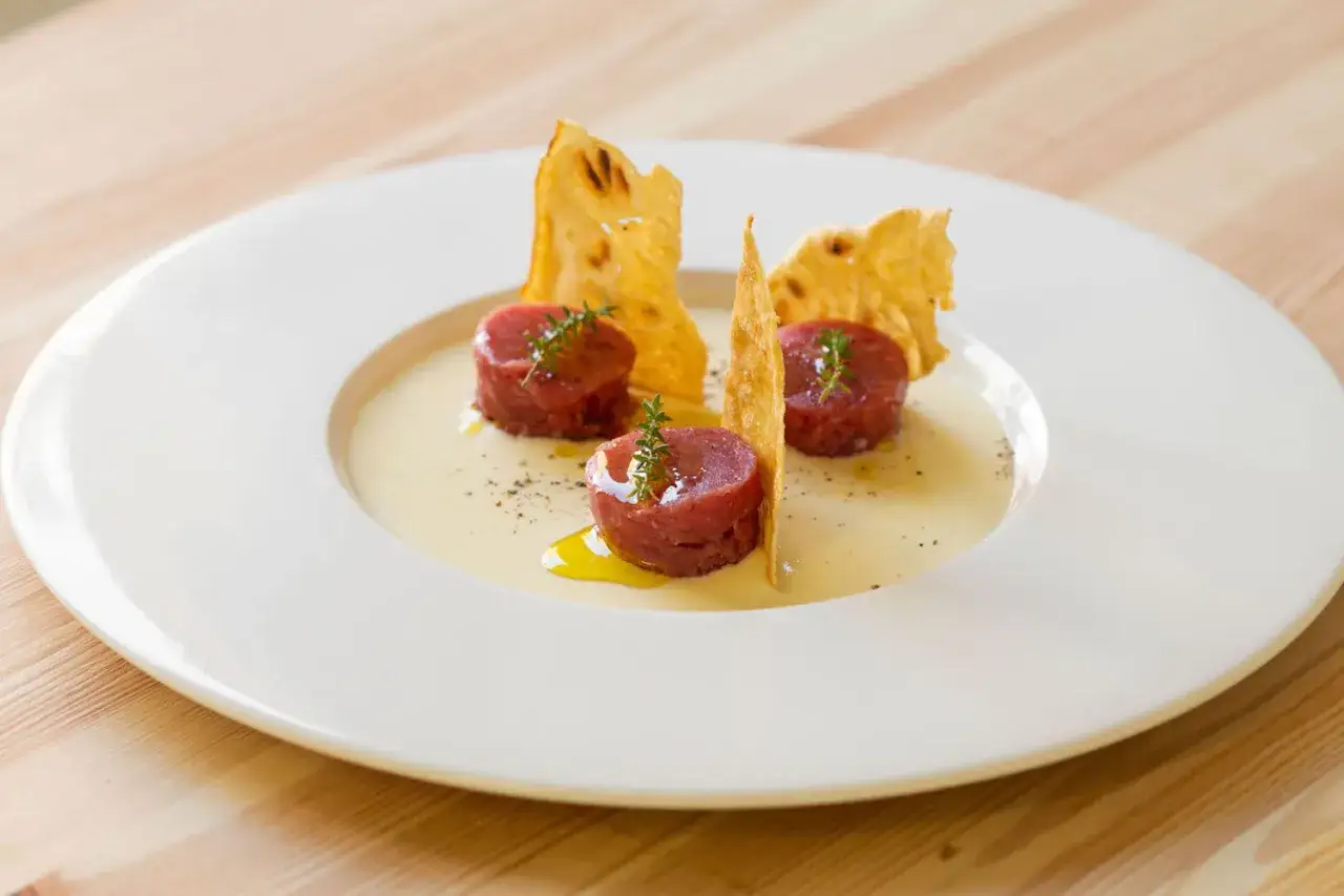 Tre cilindri di tartare di manzo, guarniti con timo, su una crema chiara e croccanti sfoglie dorate.