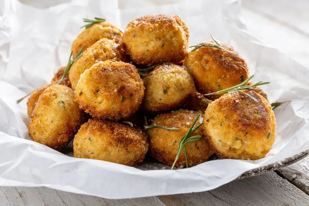 Un mucchio di polpette in bianco dorate e croccanti, guarnite con rosmarino fresco e sale grosso, servite su carta bianca.
