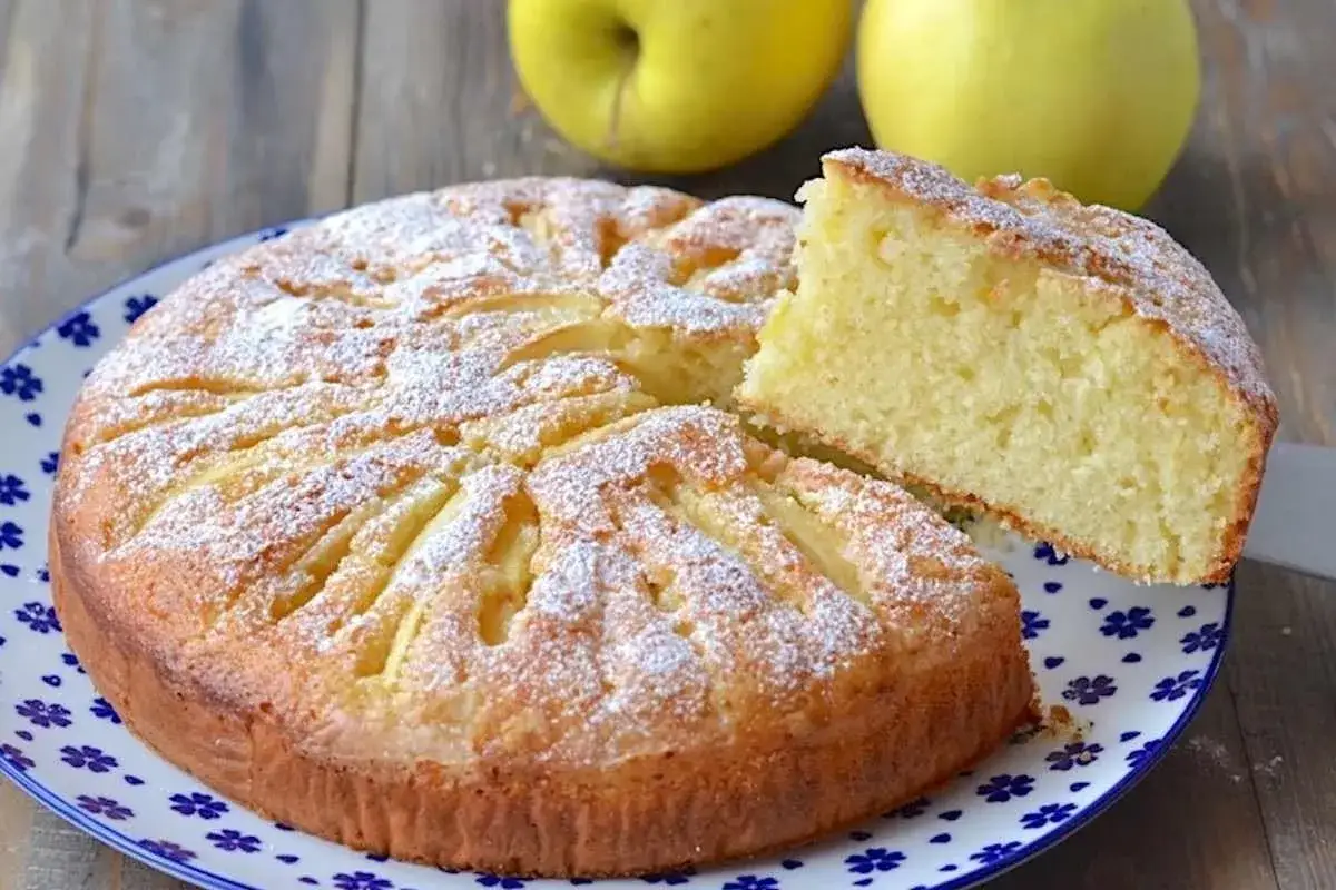 Una fetta di torta di mele con yogurt, cosparsa di zucchero a velo, su un piatto decorato. Due mele gialle sullo sfondo.