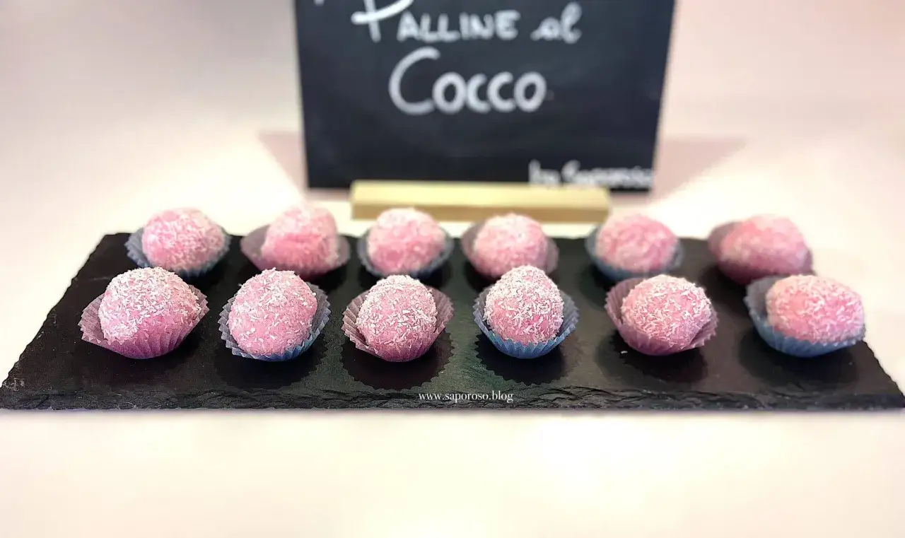 Dodici palline di cocco rosa, dolcetti invitanti ricoperti di cocco grattugiato, disposte su una lavagna scura.