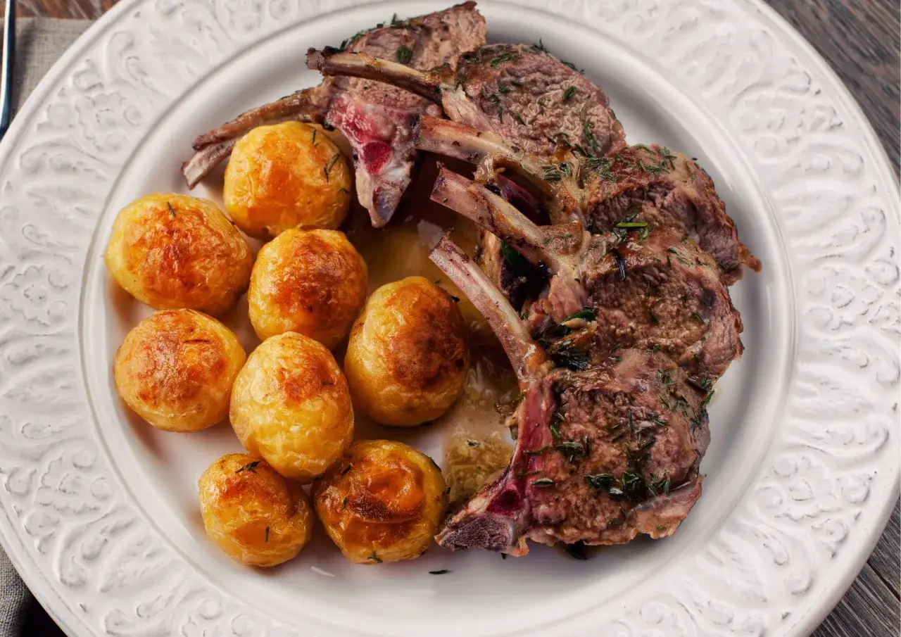 Deliziose costolette di agnello al forno, dorate e succulente, servite con patatine novelle arrosto su un piatto bianco decorato.