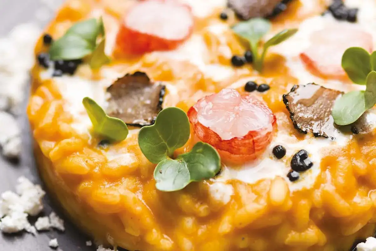 Un cremoso risotto alla zucca, guarnito con gamberi rosa, scaglie di tartufo nero, germogli verdi e briciole di formaggio.