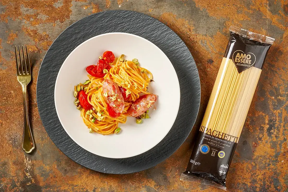 Un piatto di spaghetti con gamberetti, pomodorini e pistacchi, accanto a una confezione di spaghetti.