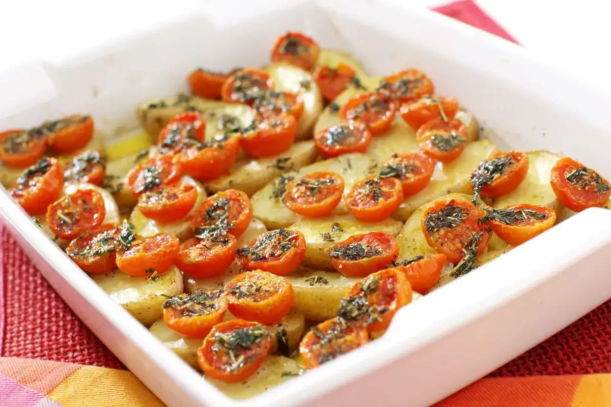 Pomodori gratinati al forno, con patate affettate e erbe aromatiche, in una teglia bianca.