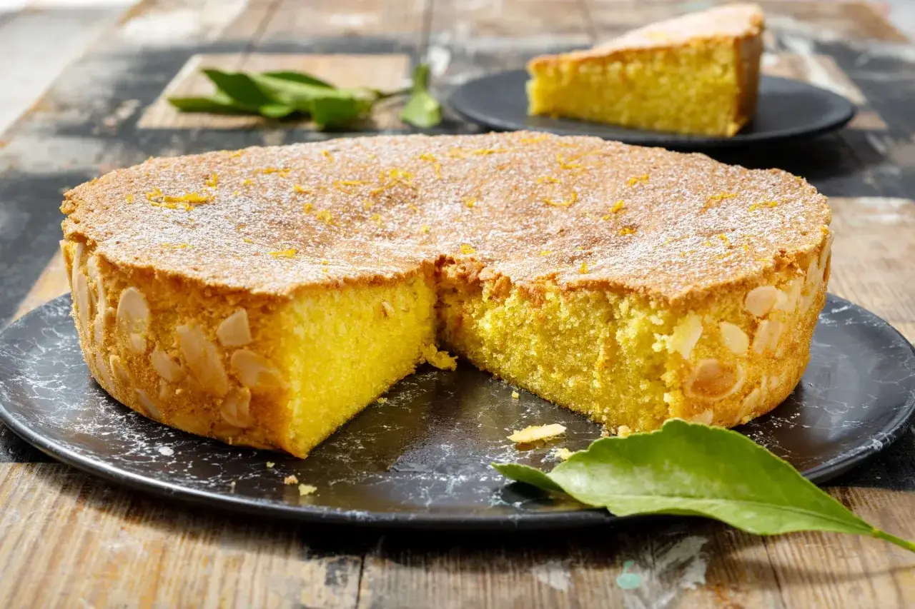 Torta soffice caprese al limone, decorata con mandorle e zucchero a velo, su un piatto scuro con foglie verdi.