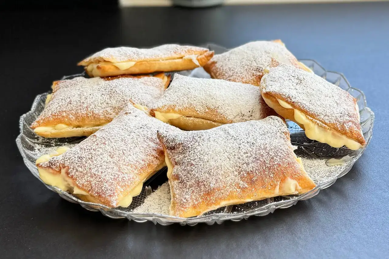 Dolci con pasta sfoglia rettangolare ripieni di crema, spolverati di zucchero a velo su un piatto di vetro.