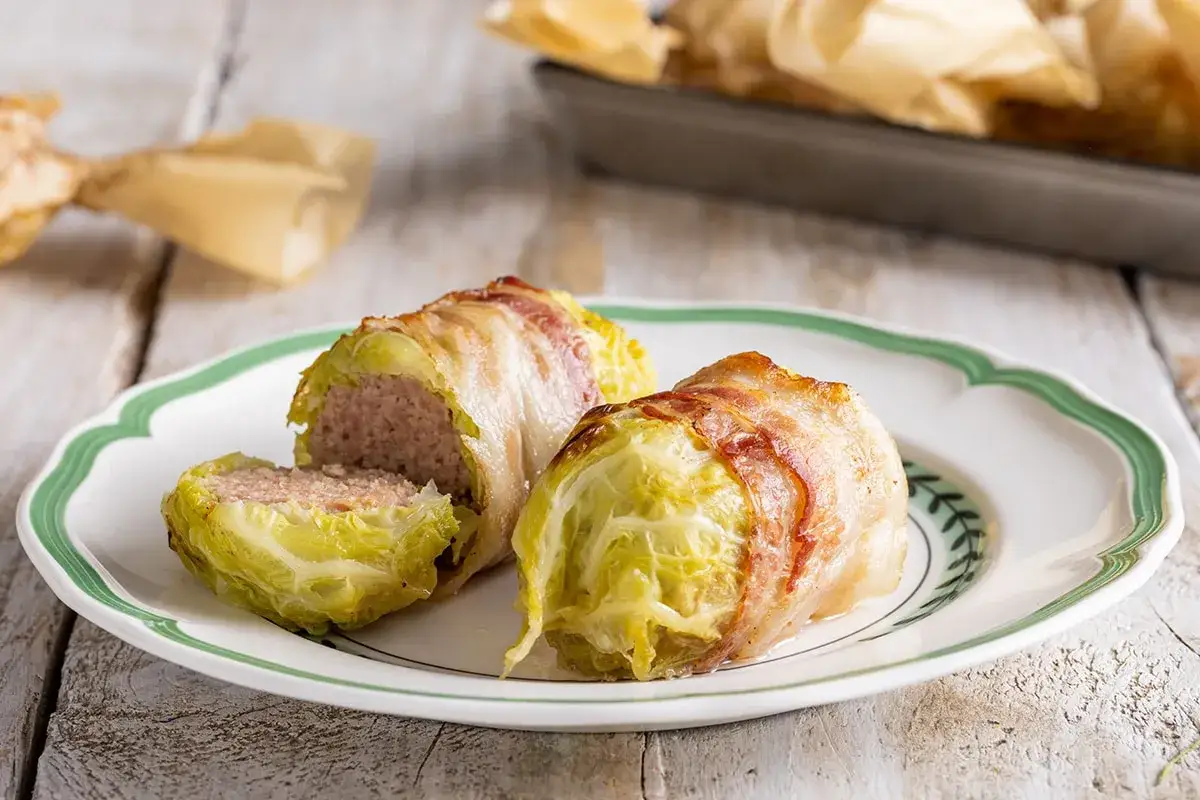 Involtini di verza con carne e pancetta, uno tagliato a metà, su un piatto bianco con bordo verde.