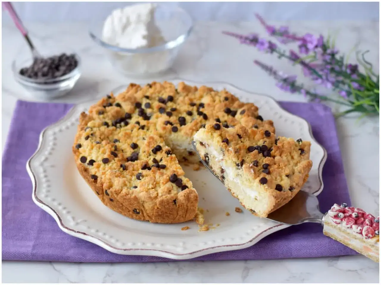 Sbriciolata ricotta e cioccolato: una fetta mostra il ripieno cremoso e le gocce di cioccolato, con ingredienti freschi sullo sfondo.