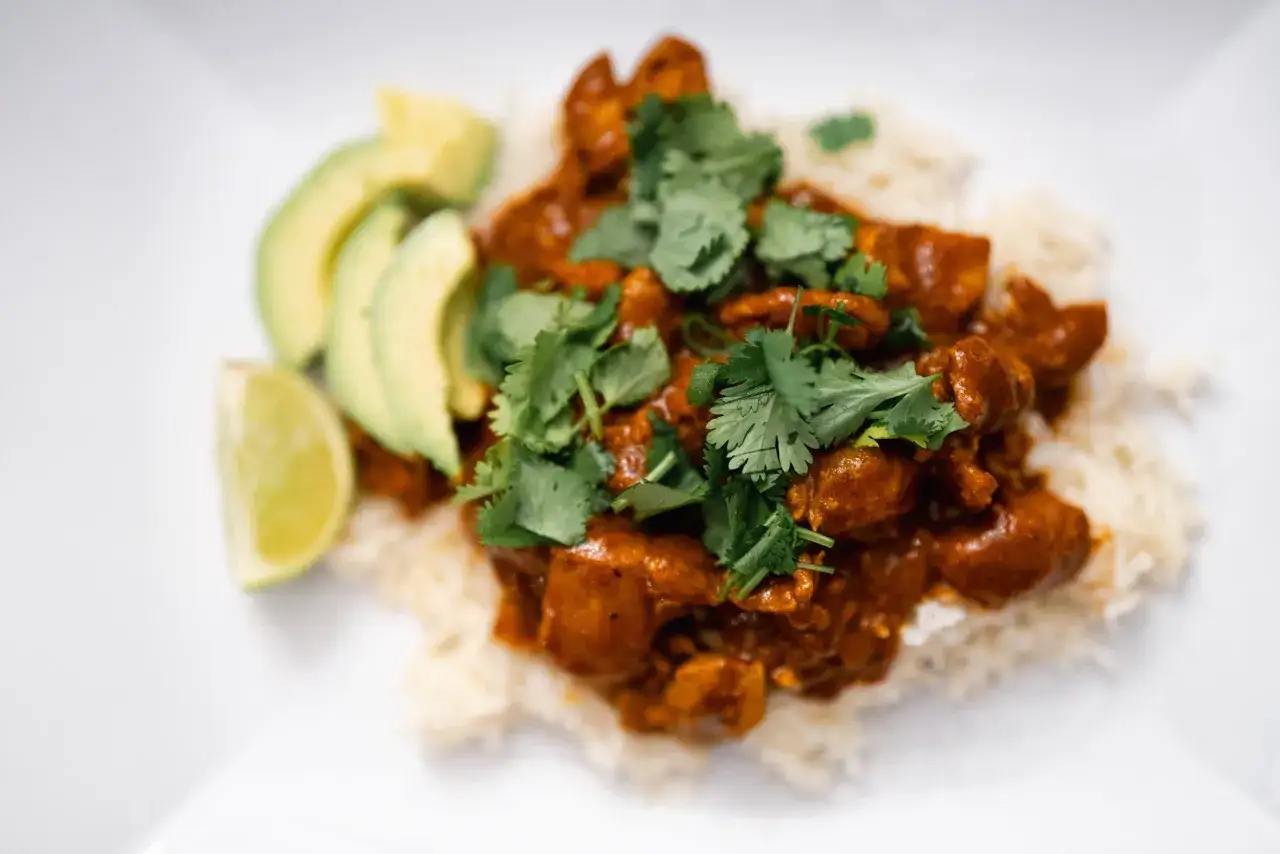 Un saporito pollo al curry con latte di cocco, servito su riso bianco, guarnito con coriandolo fresco e accompagnato da fette di avocado e lime.