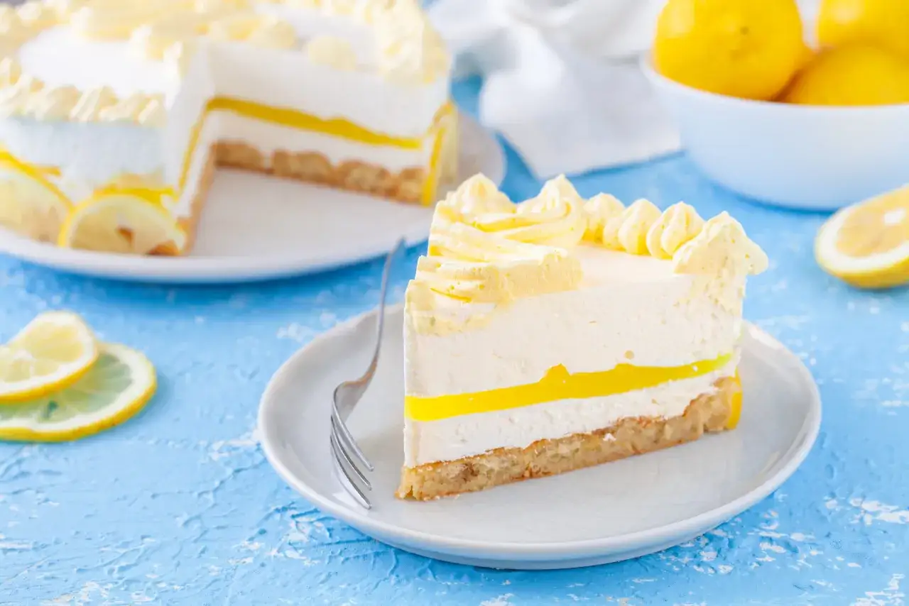 Una fetta di cheesecake al limone, con strati cremosi e una gelatina gialla brillante, guarnita con panna montata e limone.