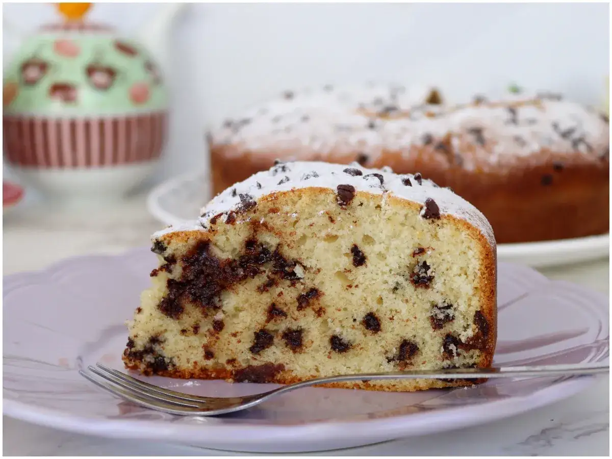 Una fetta di torta soffice con tante gocce di cioccolato, spolverata di zucchero a velo, su un piatto lilla con una forchetta.