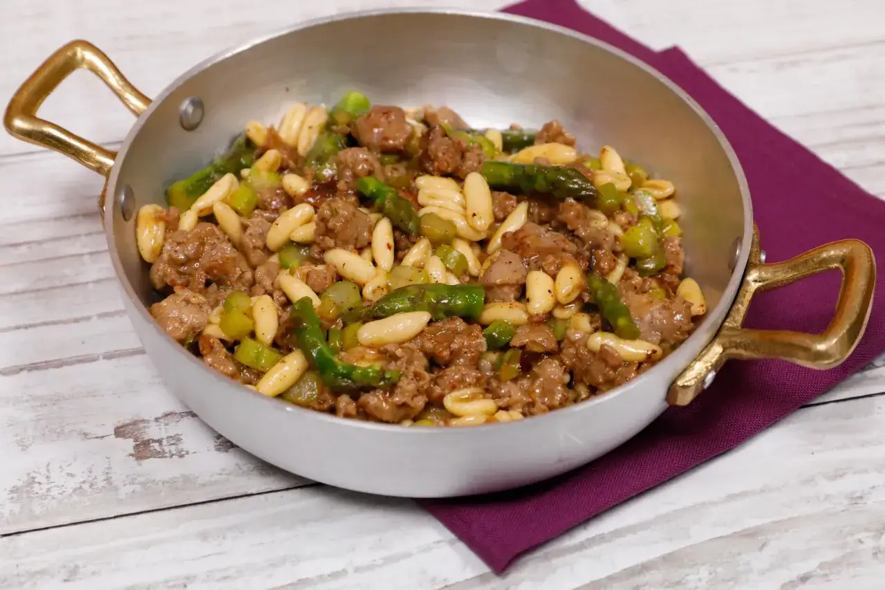 Piatto di pasta con asparagi e salsiccia, servito in una padella con manici dorati su un tovagliolo viola.