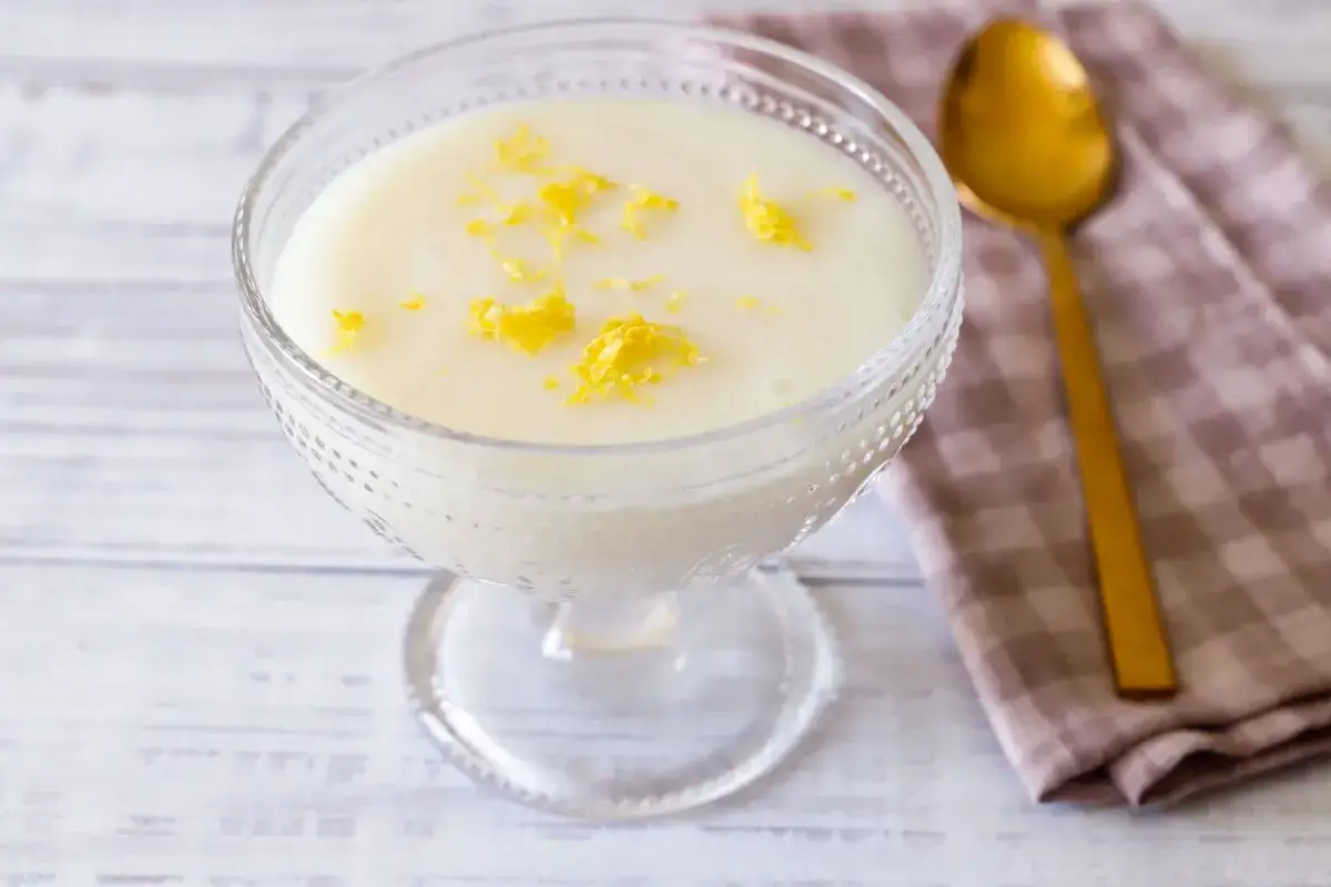 Deliziosa crema di riso in una coppa di vetro, guarnita con scorza di limone. Accanto, un cucchiaio dorato e un tovagliolo a quadretti.