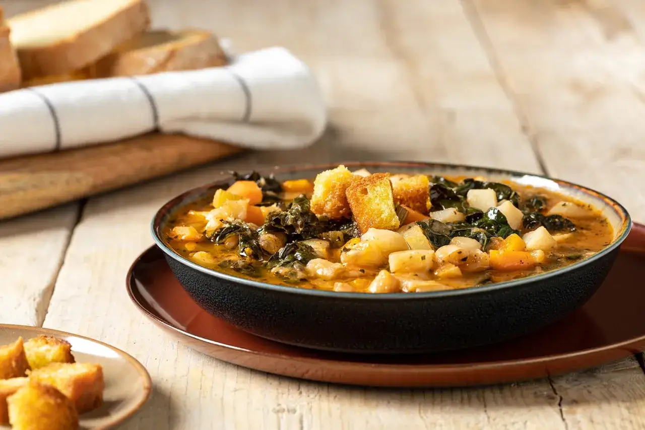 Una ciotola fumante di zuppa di cavolo nero con crostini dorati e cubetti di patate e carote. Sullo sfondo, fette di pane rustico.