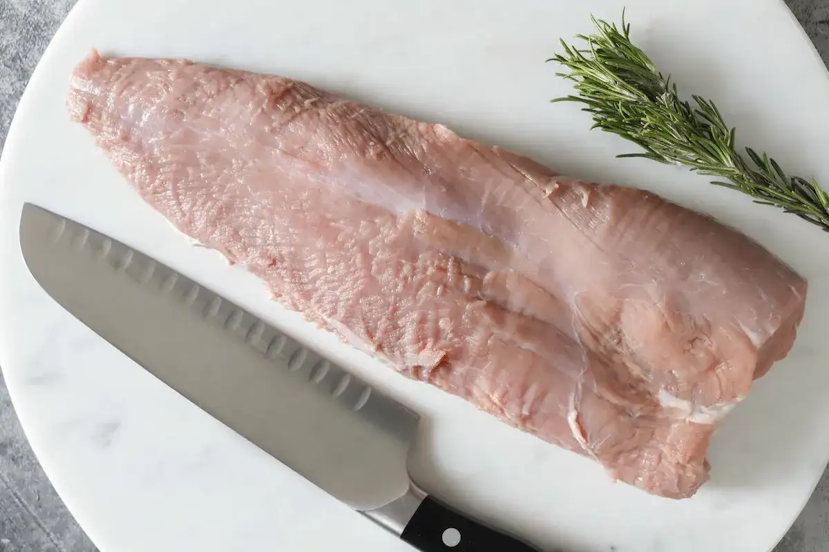 Un tenero girello di vitello pronto per essere cucinato, con un rametto di rosmarino e un coltello affilato su un tagliere di marmo.